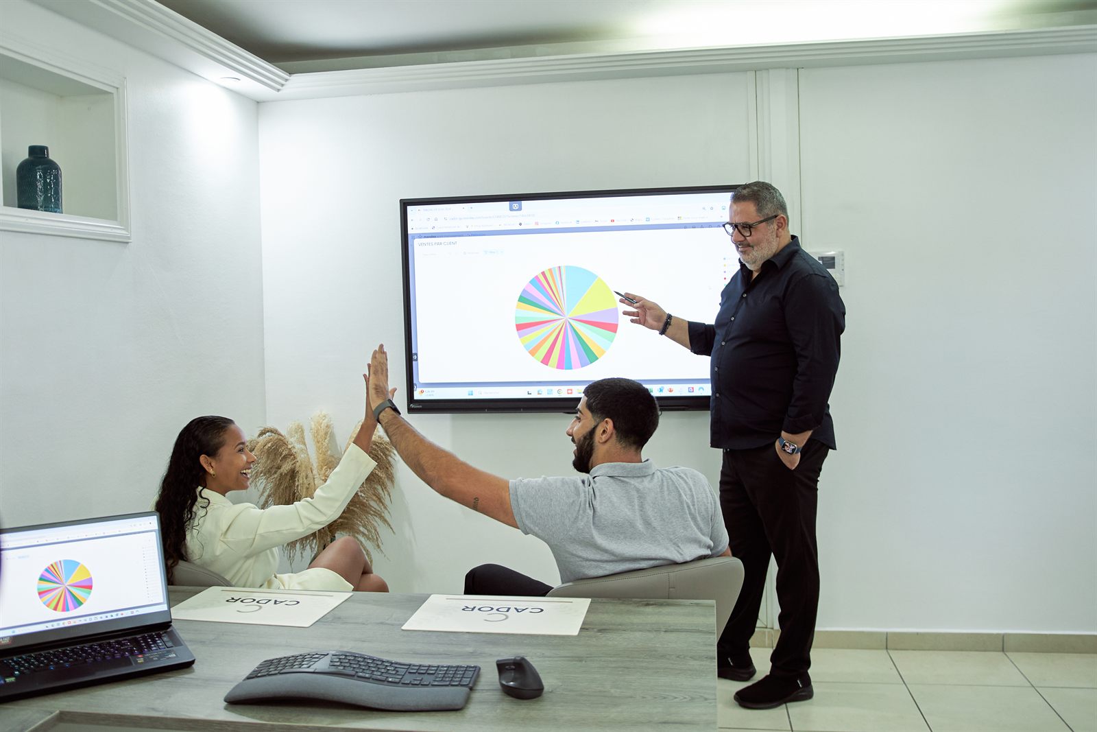 Devant un tableau de bord Monday des ventes par client, deux participants se tapent dans la main pendant que Ghassan Rizk commente le graphique